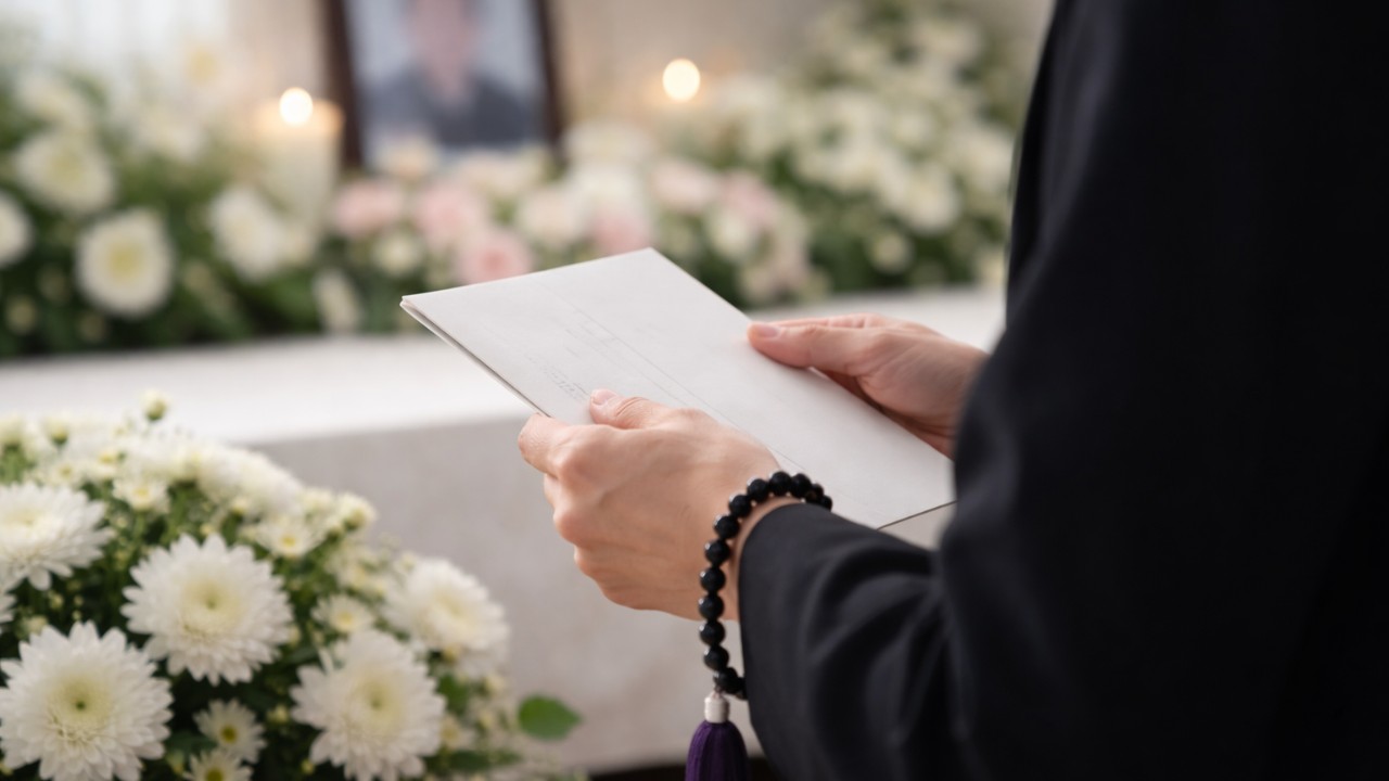 日本の葬儀を想起させる静かな式場の雰囲気、白い菊、黒い礼服の人物の手元に丁寧に持たれた手紙、